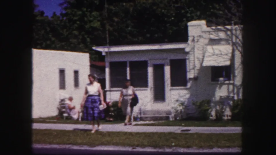 1960 Tampa frame showing two women walking past a white stucco house, with another seated figure near the doorway.