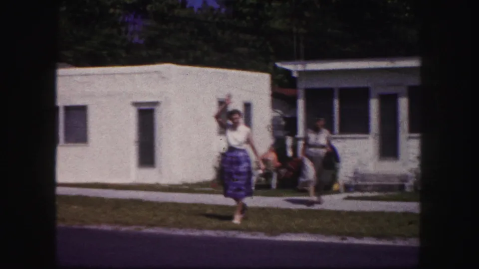 1960 Tampa home-movie frame with a woman in a white top and long patterned skirt waving as another woman follows on the sidewalk.