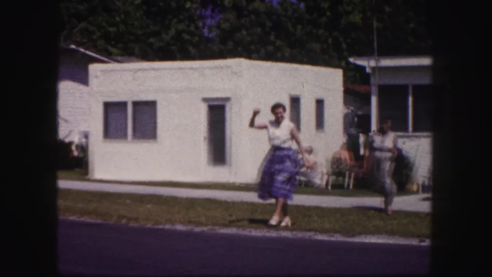 In 1960 Tampa home-movie footage, a woman waves toward the camera near a white mobile home as another woman stands farther back.