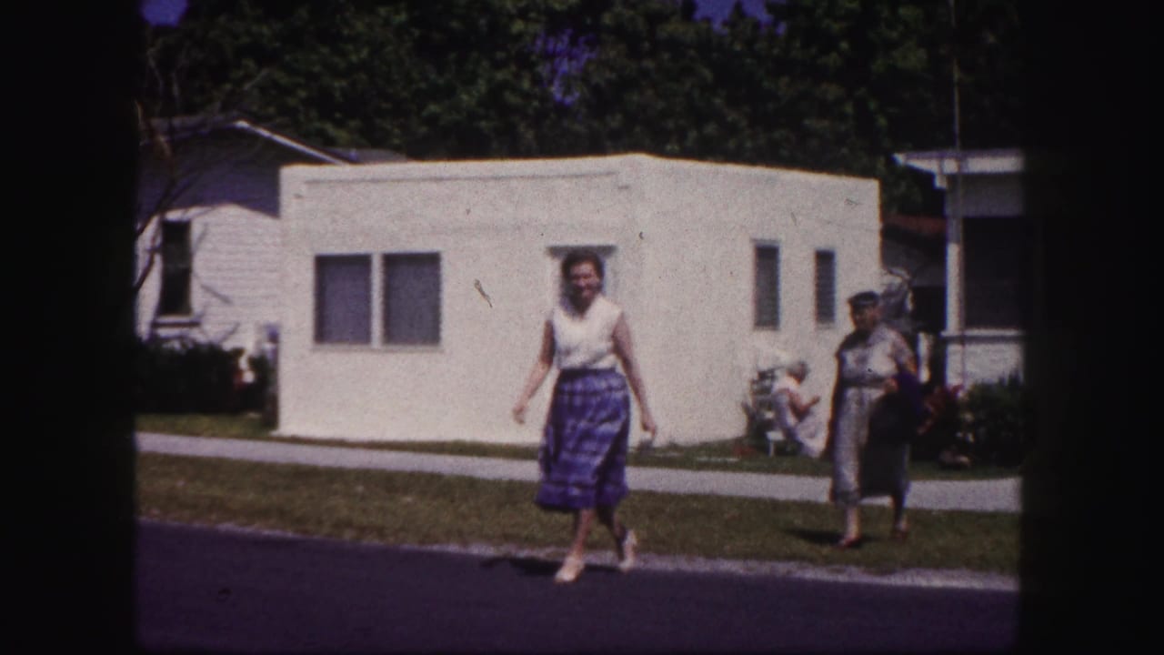 The woman in a blue plaid skirt crosses a Tampa residential street as a second woman follows near the sidewalk and mobile home.