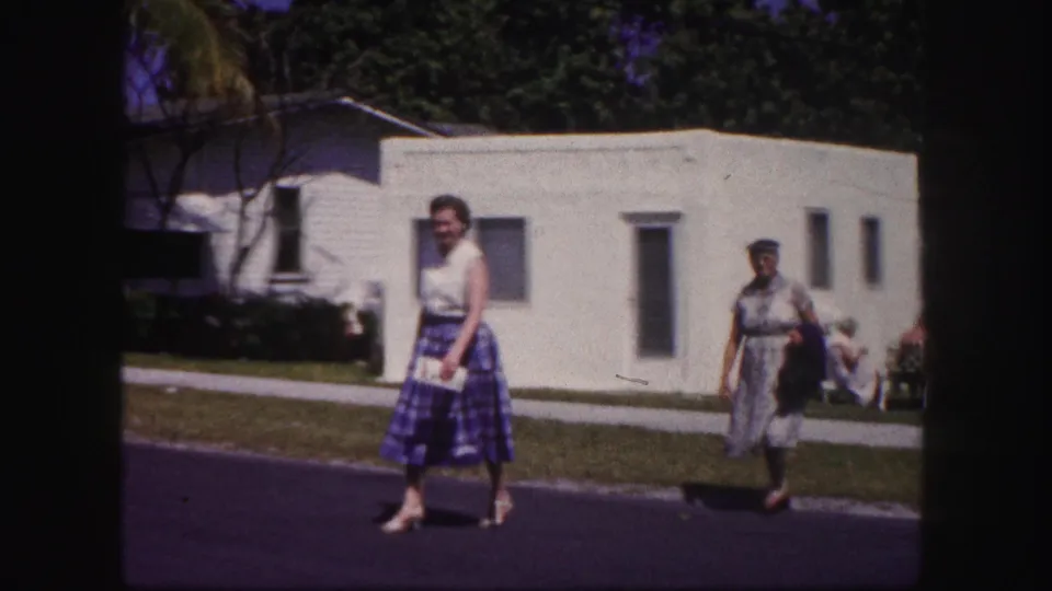 A woman in a sleeveless top and plaid skirt walks across the road, holding a small white item, with another woman near the curb behind her.