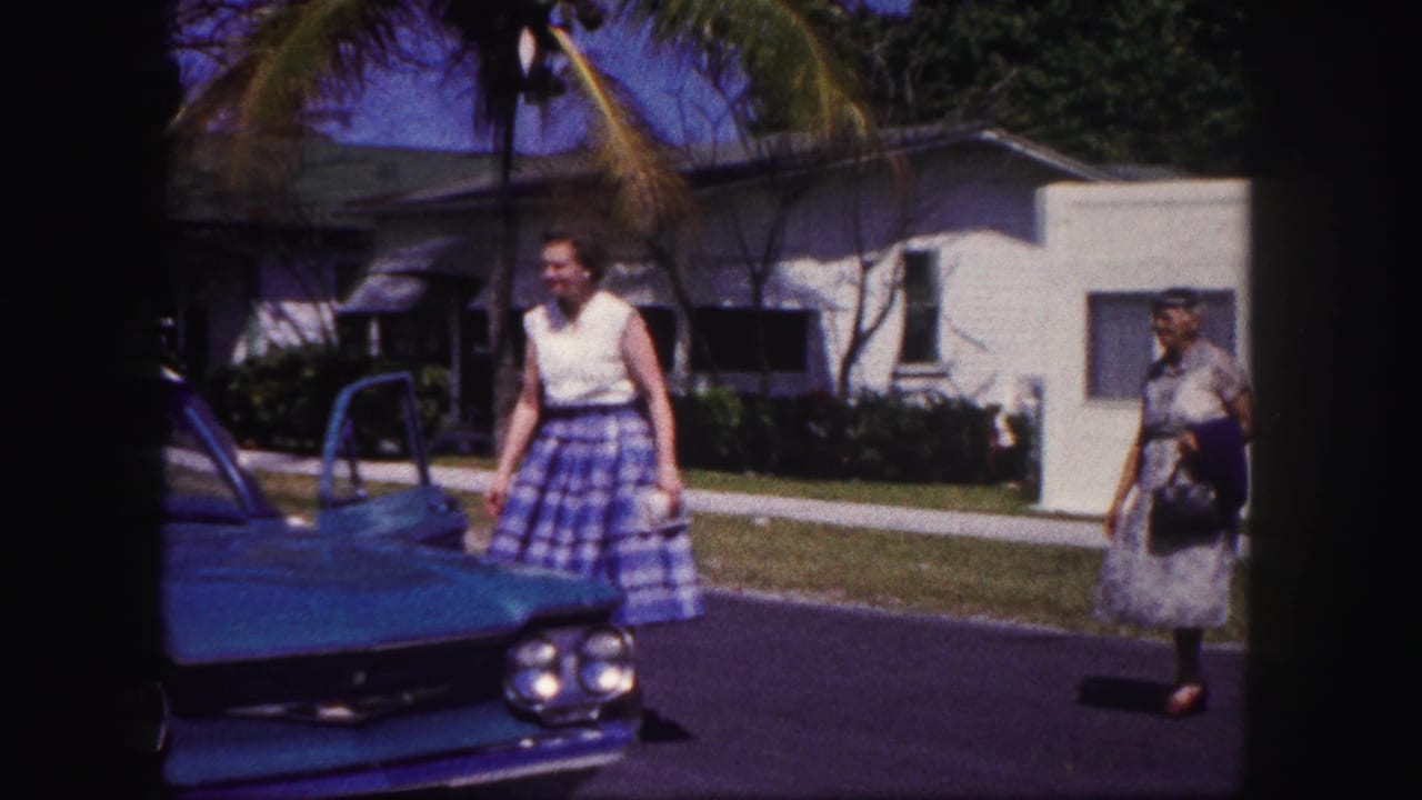 Woman in a white top and plaid skirt stands beside a turquoise car on a suburban street.