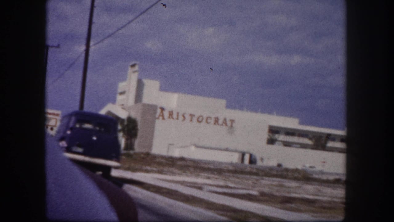 1960 Tampa frame with a blue vintage car entering the roadside view near the white ARISTOCRAT building.