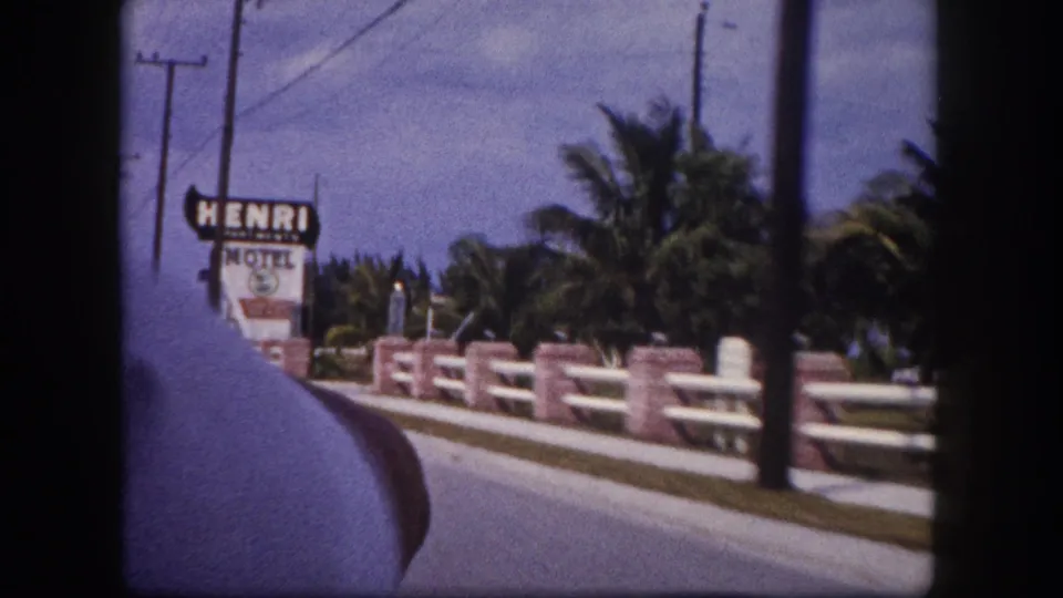1960 Tampa roadside frame showing a HENRI HOTEL sign, palm trees, and a brick-and-white guardrail from a moving vehicle.