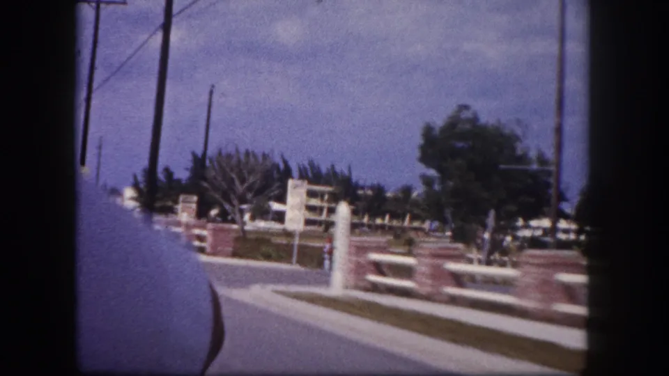 After the motel sign passes out of frame, the 1960 Tampa roadside view shows railings, palms, utility poles, sidewalk, and car interior edge.