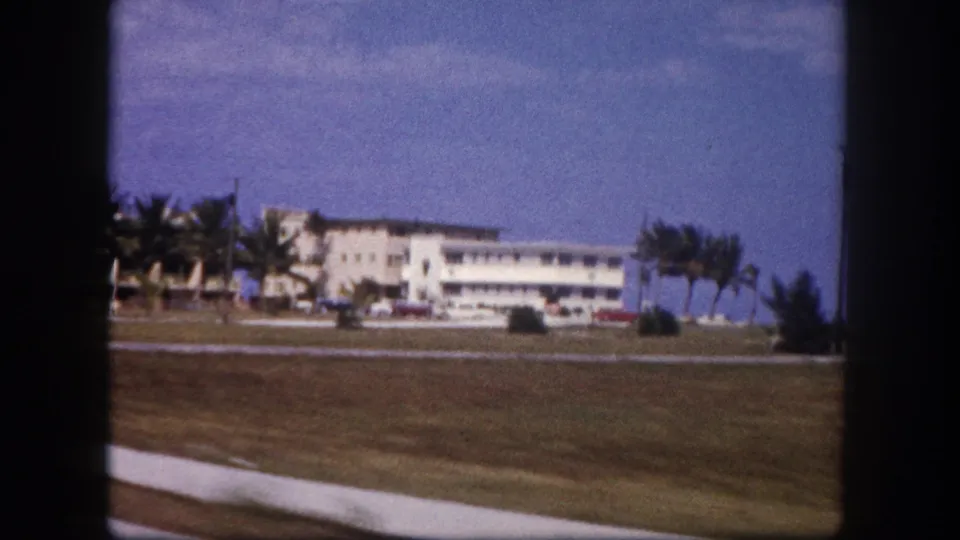 1960 Tampa passenger-view frame showing a white building, palm trees, parked cars, grass, and sidewalk under a bright sky.