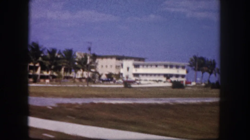 1960 Tampa road-view frame with a white building, palms, grass, sidewalk, parked cars, and visible 8mm gate edges.