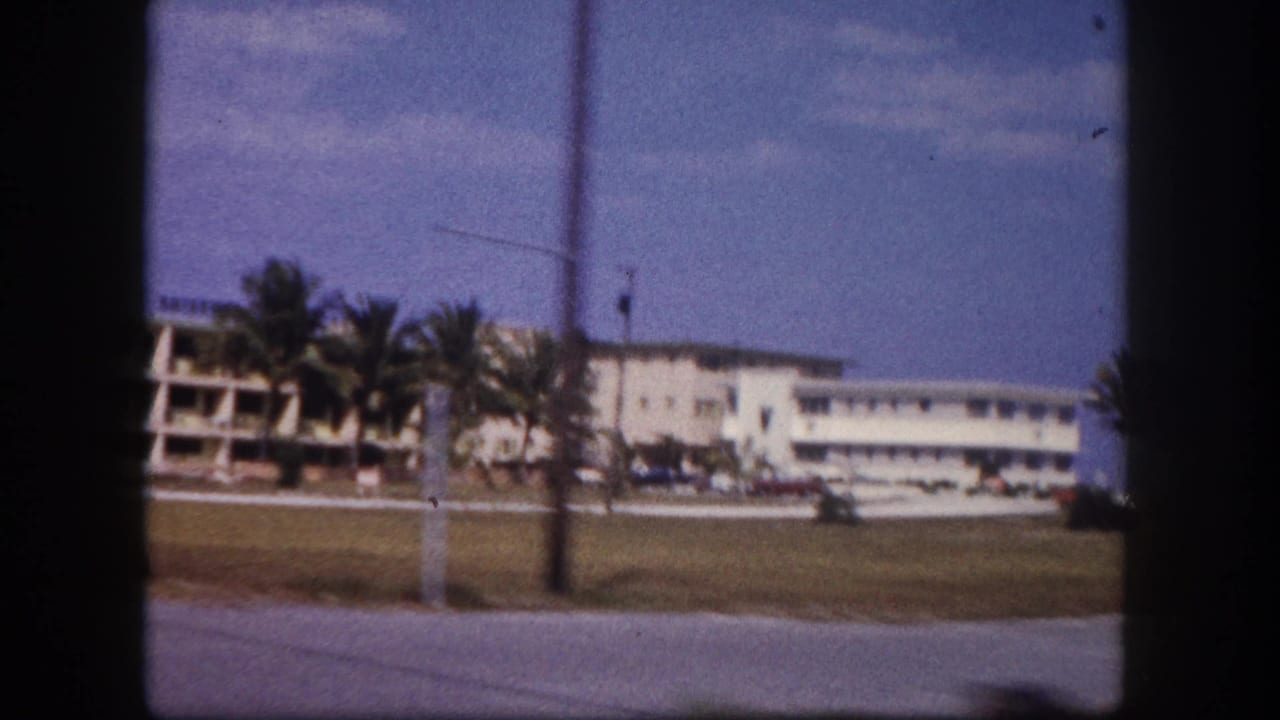 1960 Tampa home-movie frame showing a passing white building complex, palm trees, lawn, roadway edge, and blue sky.