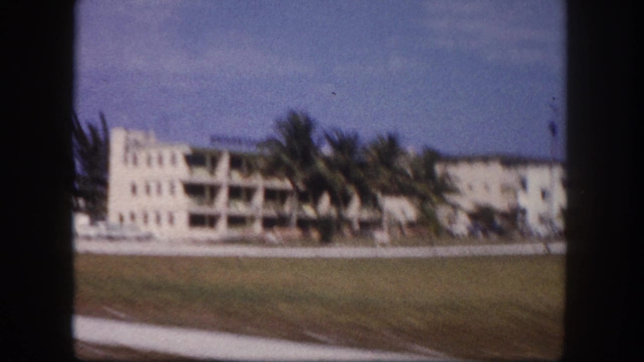 1960 Tampa roadside frame with a white multi-story building, balcony lines, palm trees, grass, sidewalk, and bright sky.
