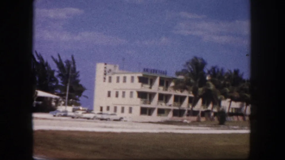 1960 Tampa frame showing a closer roadside view of a white building, palm trees, parked cars, lawn, and open sky.