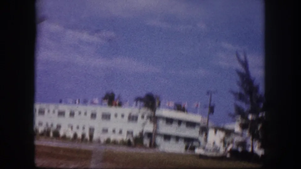 1960 Tampa home-movie frame with a blurred white roadside building, roofline flags, palm tree, lawn, utility pole, and sky.