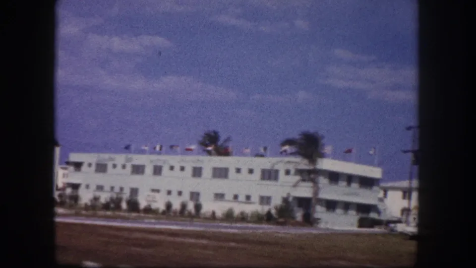 1960 Tampa home-movie view of a white motel frontage across a road, with roof flags, palm trees, and classic cars nearby.