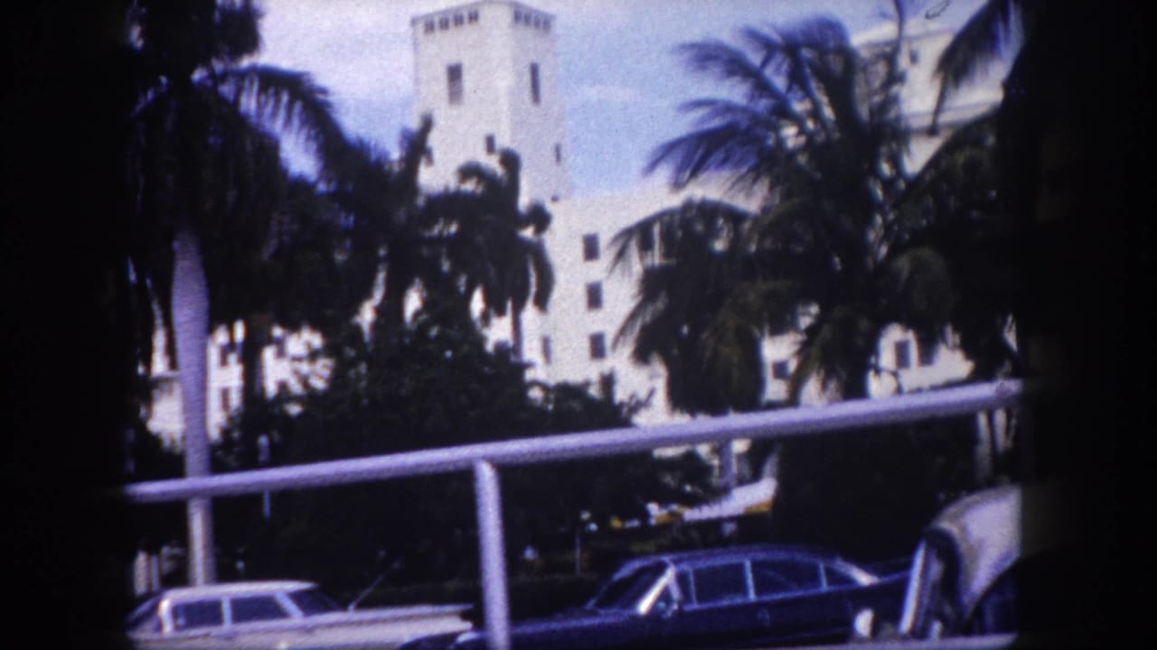 White towered building behind palms with parked cars and a railing in the foreground.