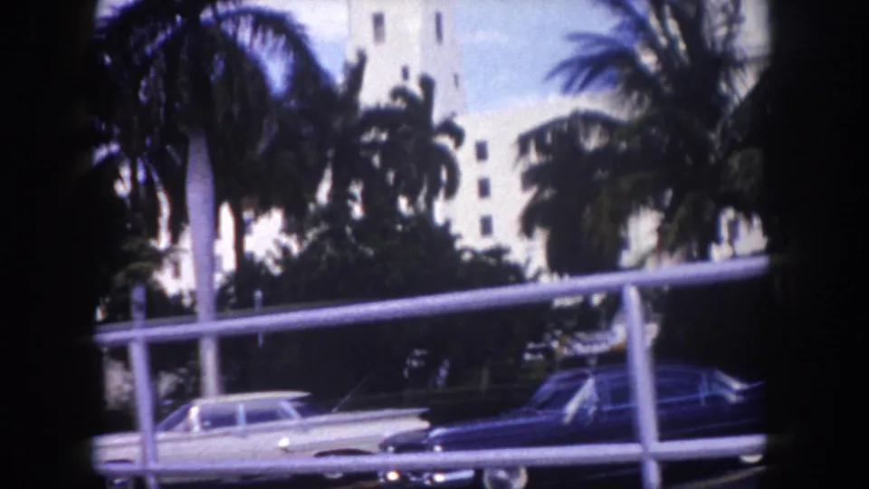 1960 Tampa frame showing palms, a white mid-century building, a light sedan, and a dark sedan along the curb.