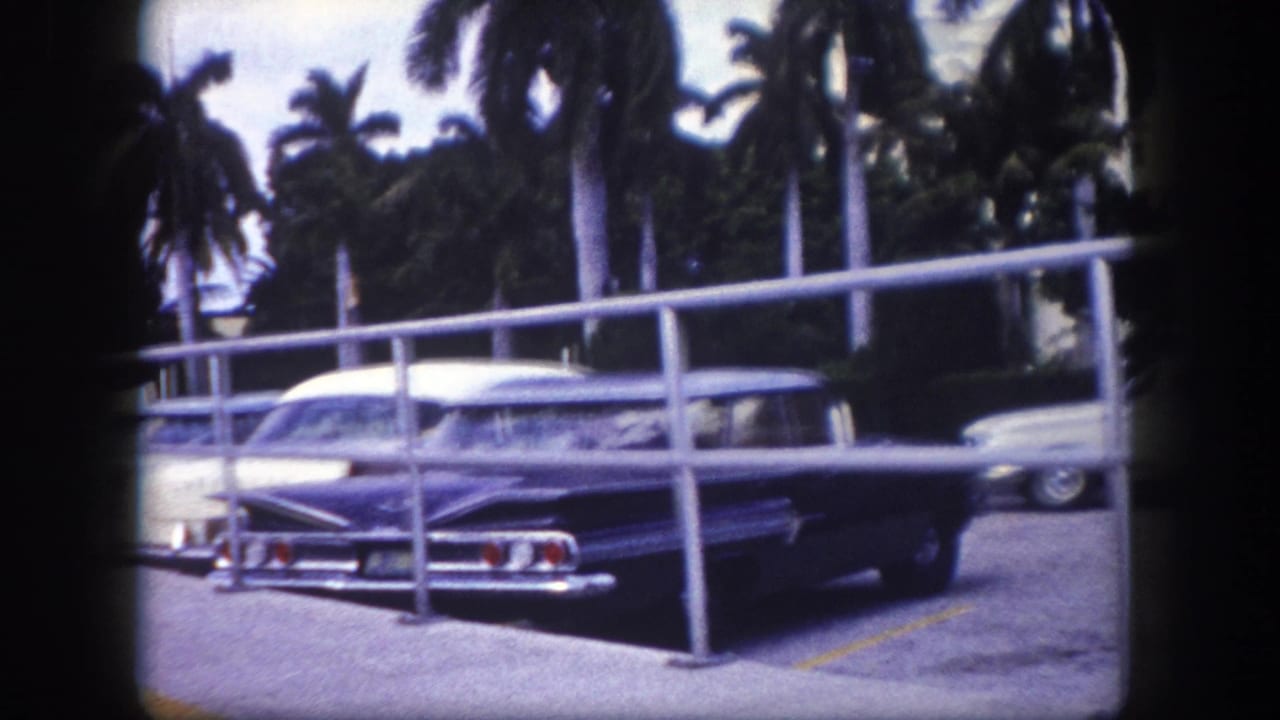 1960 Tampa home-movie frame showing a dark blue mid-century car parked behind railings near palm trees.