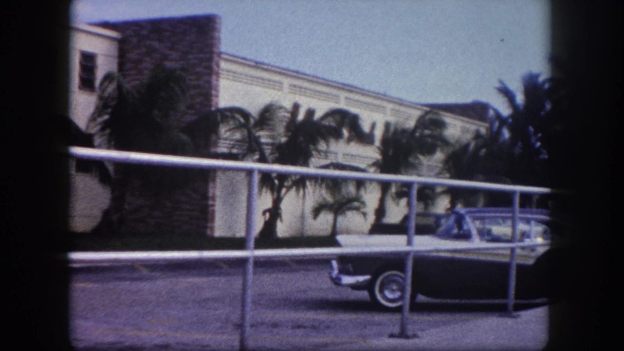 1960 Tampa 8mm frame showing a two-tone sedan parked outside a palm-lined hotel or motel exterior behind a metal railing.