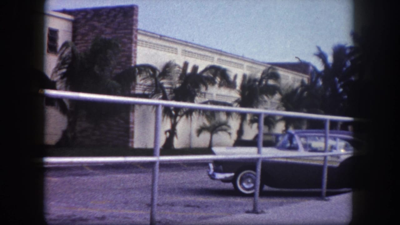 1960 Tampa frame with a stationary sedan in front of palms and a low-rise lodging-style building, partly obscured by a railing.