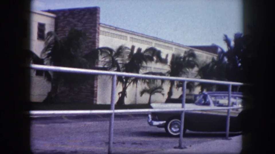 1960 Tampa 8mm view of a parked sedan and palm trees outside a low-rise hotel or motel-like building from a raised walkway.
