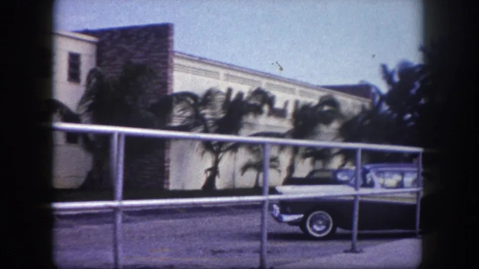 1960 Tampa frame showing the front portion of a parked sedan beneath a metal railing, with palm trees and a low-rise exterior behind it.