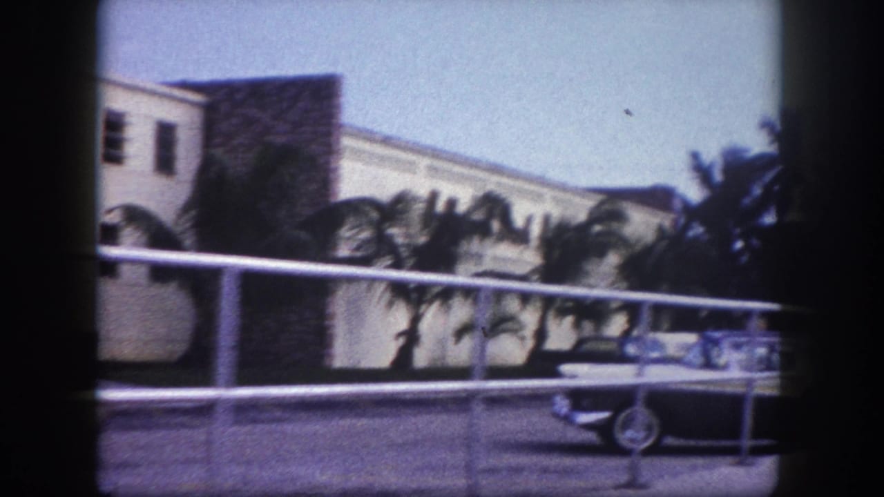 1960 Tampa street view with a classic car near a railing, palms, and a light-colored building in soft 8mm color.
