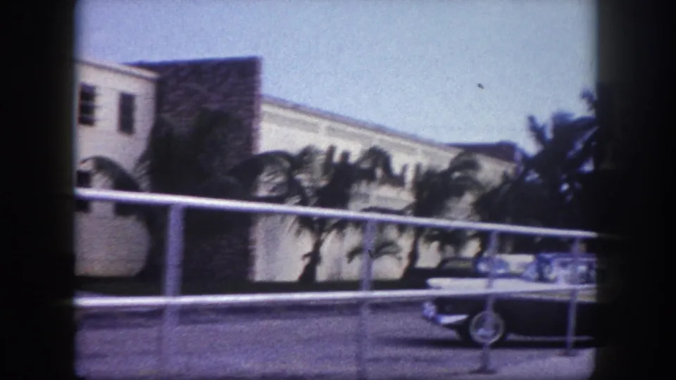 1960 Tampa street view with a classic car near a railing, palms, and a light-colored building in soft 8mm color.