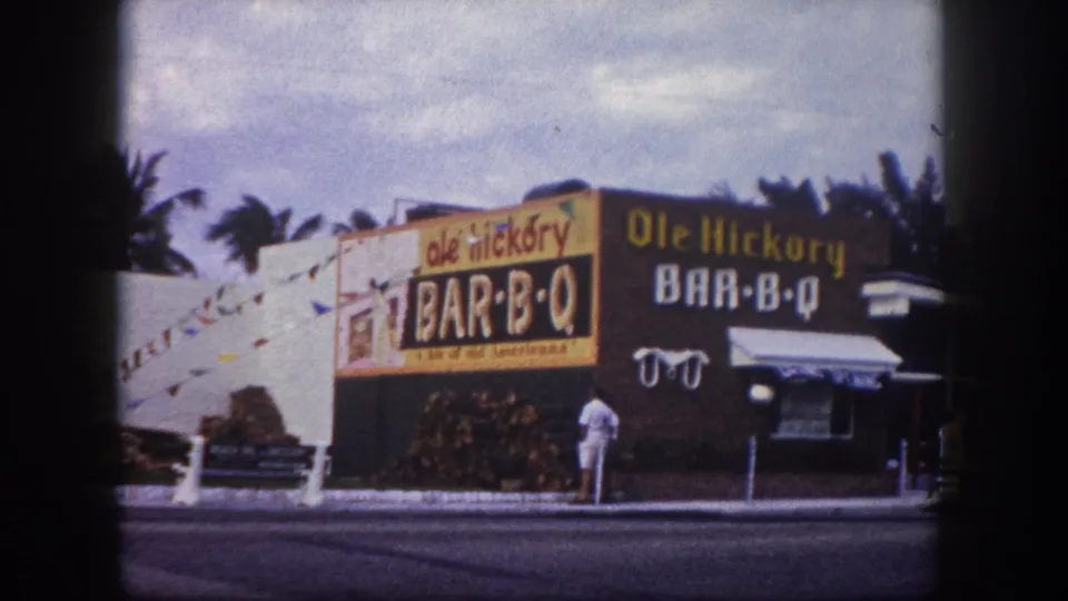 Ole Hickory Bar-B-Q exterior in Tampa, 1960, with large signage, stacked firewood, palms, sidewalk, and a visible pedestrian.