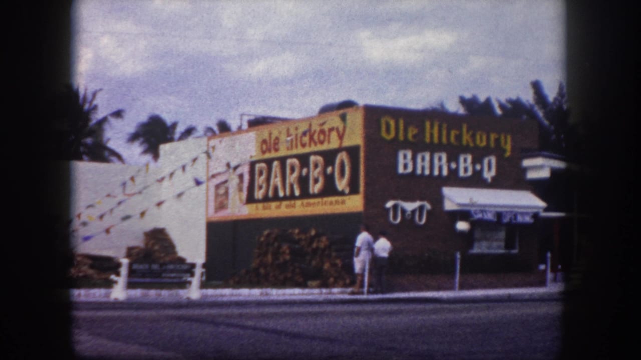 1960 Tampa home-movie frame showing the Ole Hickory Bar-B-Q facade, yellow and brown signs, stacked wood, palms, and two adults on the sidewalk.