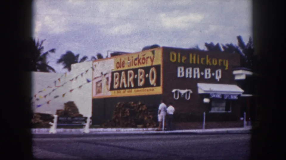 1960 Tampa home-movie frame showing the Ole Hickory Bar-B-Q facade, yellow and brown signs, stacked wood, palms, and two adults on the sidewalk.