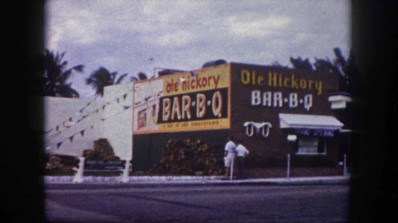 1960 Tampa frame of the Ole Hickory Bar-B-Q building, with bold exterior signage, firewood piles, palms, awning, and adults by the doorway.