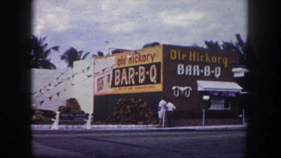 1960 Tampa frame of the Ole Hickory Bar-B-Q building, with bold exterior signage, firewood piles, palms, awning, and adults by the doorway.