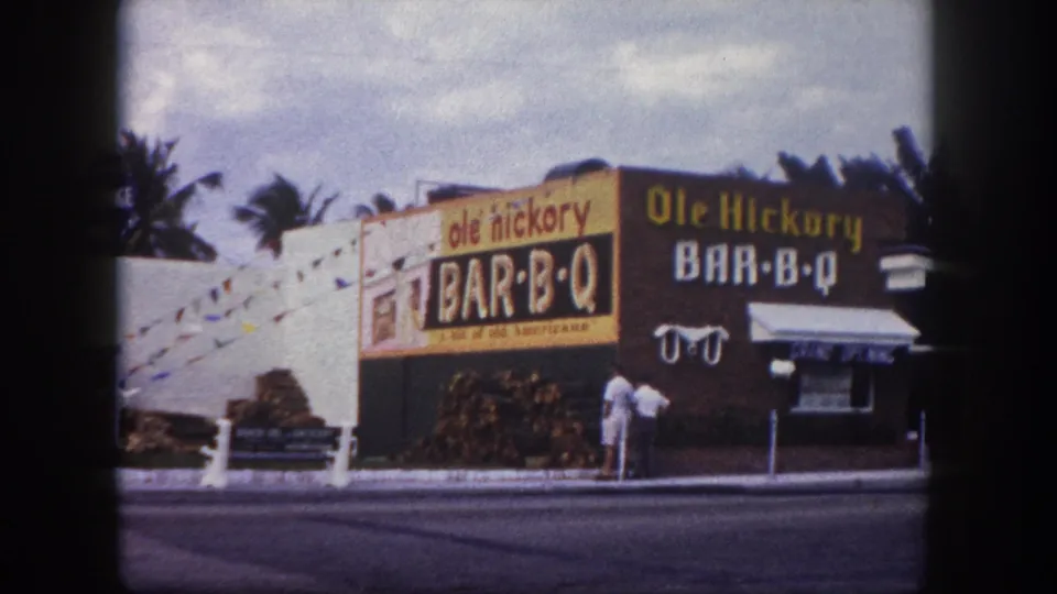 1960 Tampa 8mm frame holding on the Ole Hickory Bar-B-Q exterior, showing signage, stacked wood, palms, sidewalk, and adults near the front.