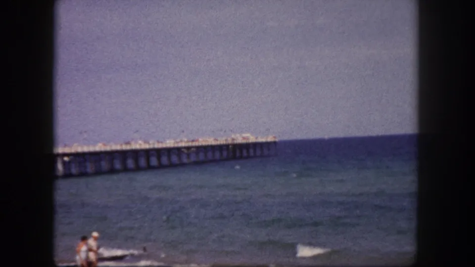 A man and woman appear near the Tampa shoreline while the long pier remains in the background on 1960 8mm footage.