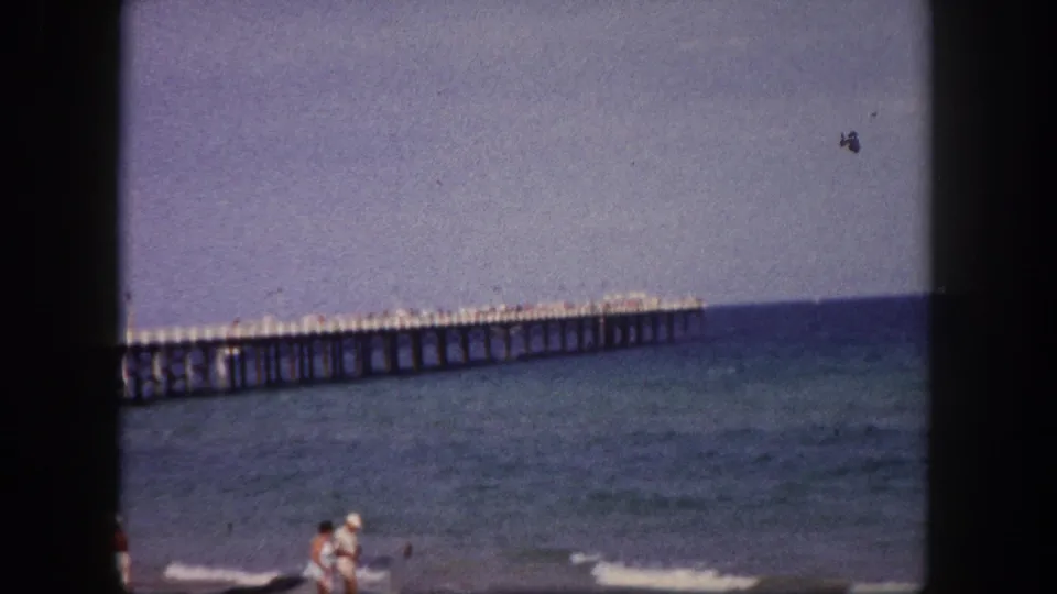 1960 Tampa beach frame showing a couple walking along the shoreline, with surf and a long pier behind them.