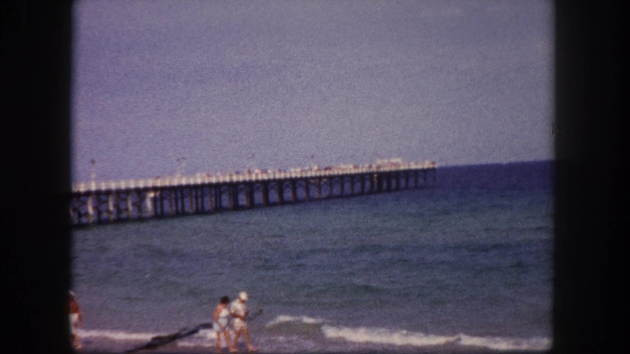 1960 Tampa beach home-movie frame showing a couple near the surf with a long ocean pier in the background.