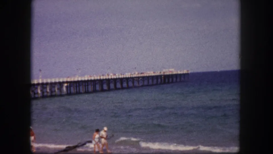 1960 Tampa beach home-movie frame showing a couple near the surf with a long ocean pier in the background.