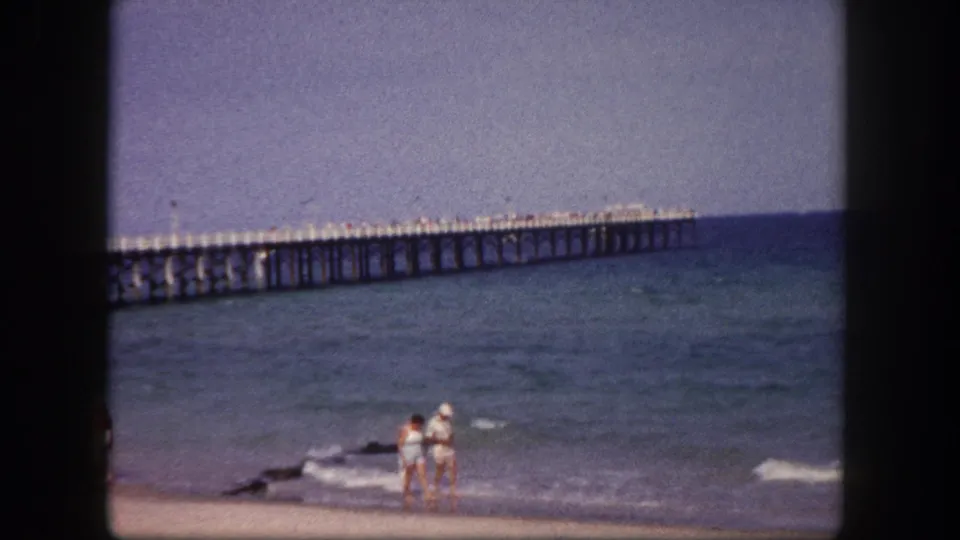 1960 Tampa shoreline view with a couple in light beachwear near small waves and a long pier in the distance.