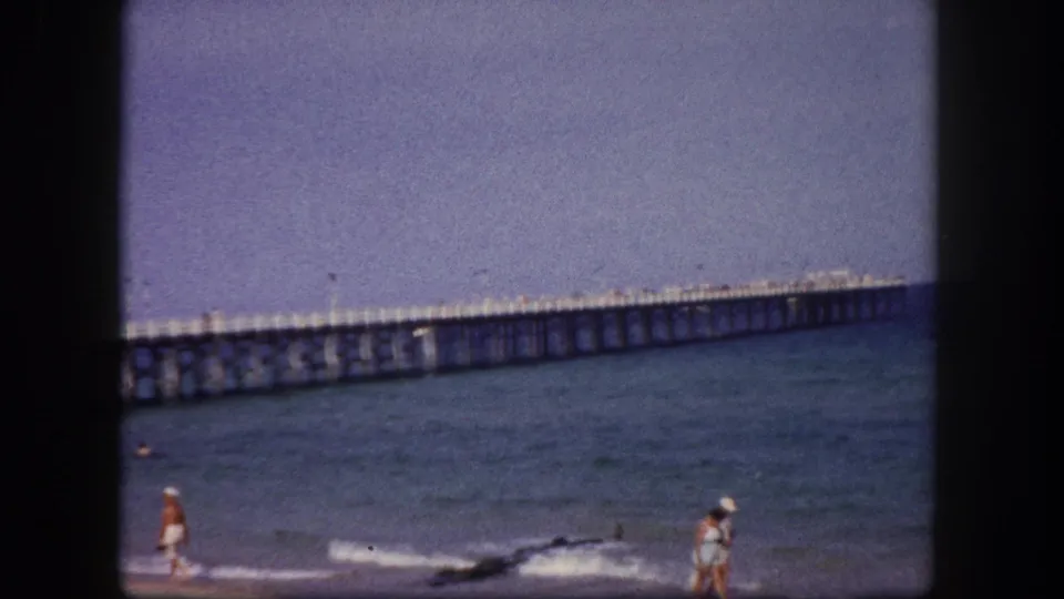 1960 Tampa beach home-movie frame with distant beachgoers, low surf, and a long ocean pier behind them.
