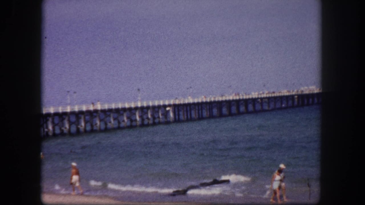 Beachgoers near small surf with a long wooden pier stretching across the water.