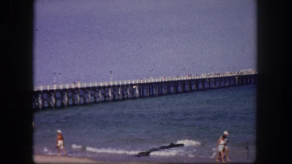 1960 Tampa-area beach view with walkers near the shoreline and a long wooden fishing pier crossing the background.