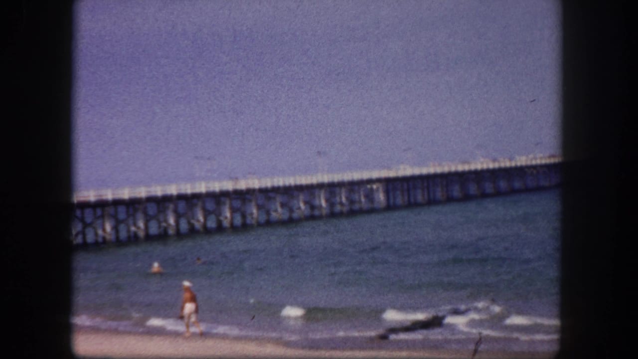 A person walks along the surf while a long pier spans the water behind them.