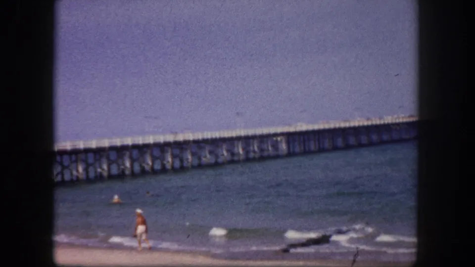 A beachgoer walks at the edge of the surf beneath a long wooden pier in 1960 Tampa-area home-movie footage.