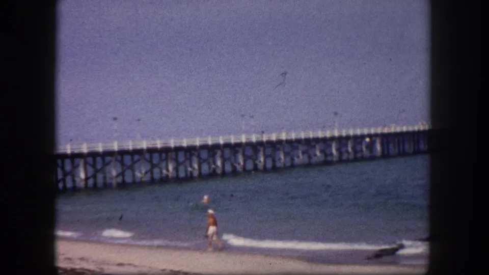 Tampa-area beach scene showing a figure near the waterline, low surf, and the wooden pier across the horizon.