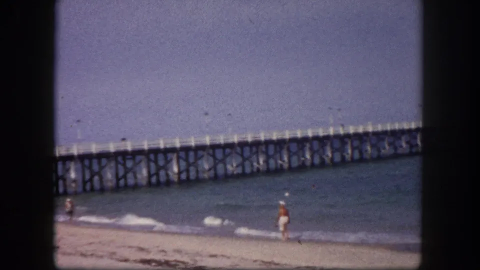 End frame of the 1960 Tampa-area beach clip with a walker near the surf and the long wooden pier visible beyond.