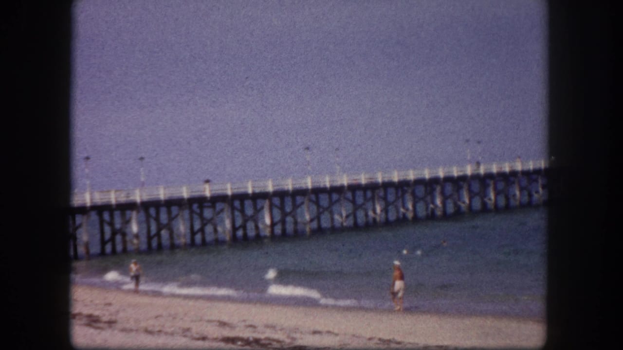 Wide 8mm shot of a sandy beach with a timber trestle pier, distant bathers, and a blue-shifted sky — Tampa, Florida, circa 1960.