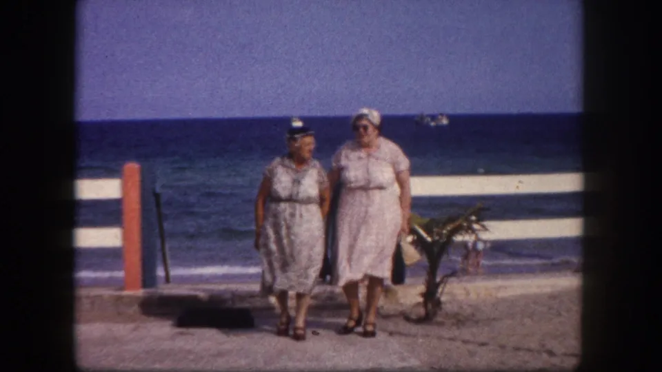 Close continuation of two women in day dresses and white sun hats on a beachside promenade, ocean horizon behind them, Tampa, Florida, circa 1960.