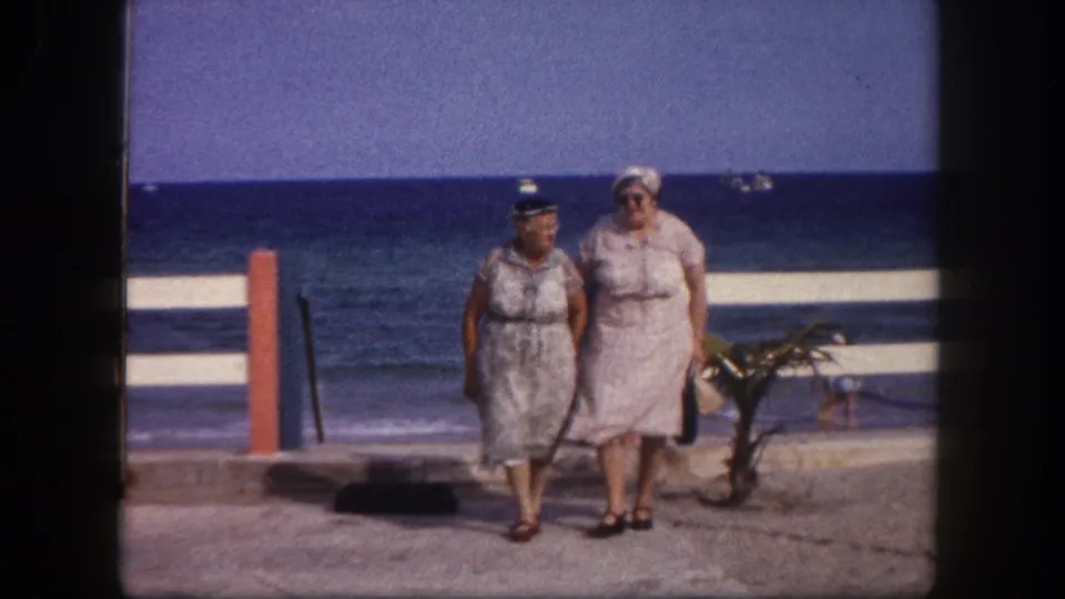 Two adult women in belted summer dresses and white sun hats walk a beachfront promenade; metadata identifies Tampa, Florida, circa 1960.