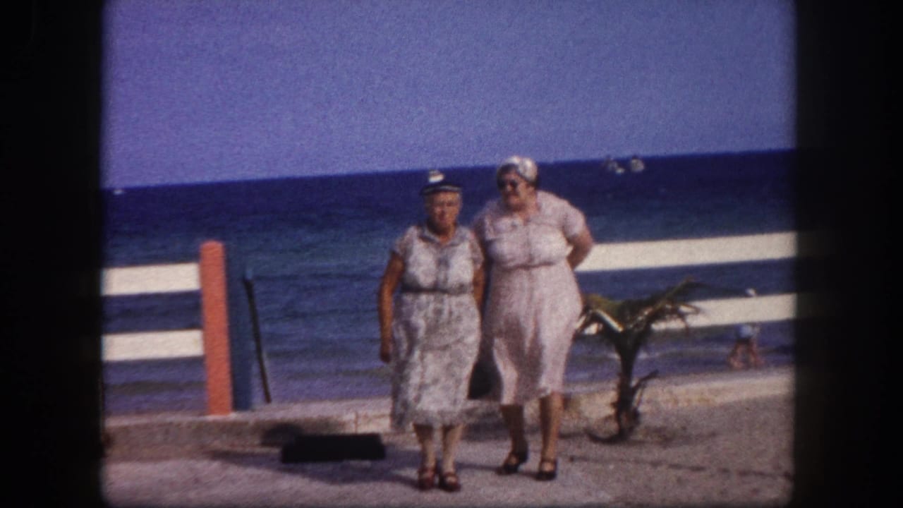 Two adult women in white belted summer dresses and sun hats walk a beachfront promenade; horizontal white railing and blue sea behind, Tampa, Florida, circa 1960.