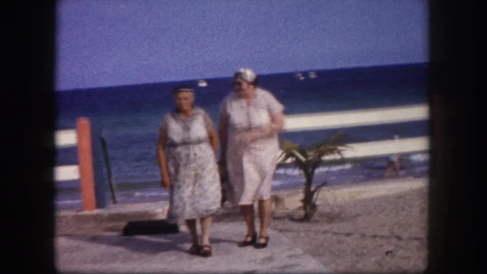 Two adult women in belted summer dresses and white hats stride along a beachside promenade; railing at left, small palm and blue ocean in background, Tampa, Florida, circa 1960.