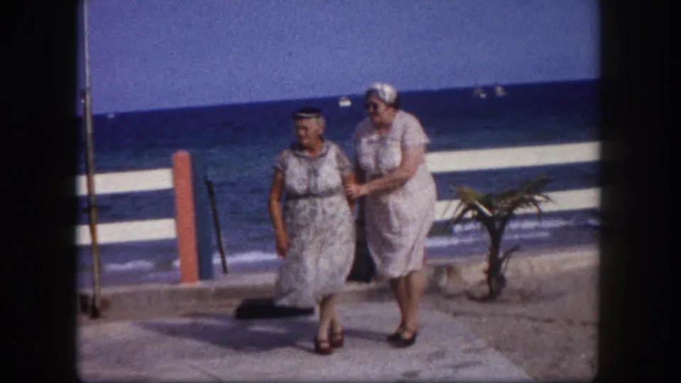 Two adult women in white belted dresses and sun hats walk closely together on a beachfront promenade; reddish-orange railing posts and calm blue ocean visible, Tampa, Florida, circa 1960.