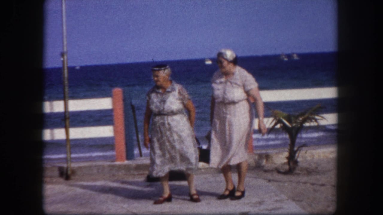 Two women in light patterned dresses and headscarves walking a concrete seaside promenade with red bollards and blue ocean behind them.
